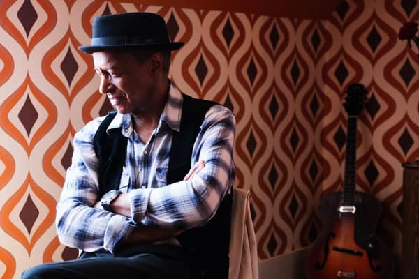 A man in a hat sitting on a chair in front of an orange wall.