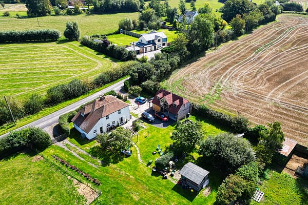 Aerial photograph of a rural area showcasing two houses with attached gardens, a few parked cars, green fields, and harvested farmland — drone property photography by Ben Bowles