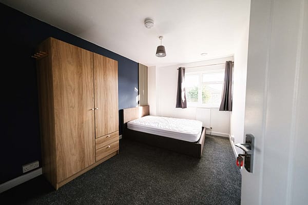 A tidy bedroom with a double bed, wooden wardrobe, and curtained window. The room has dark carpeting and white walls. A single ceiling light hangs in the center, a beautiful property image - property photography by Ben