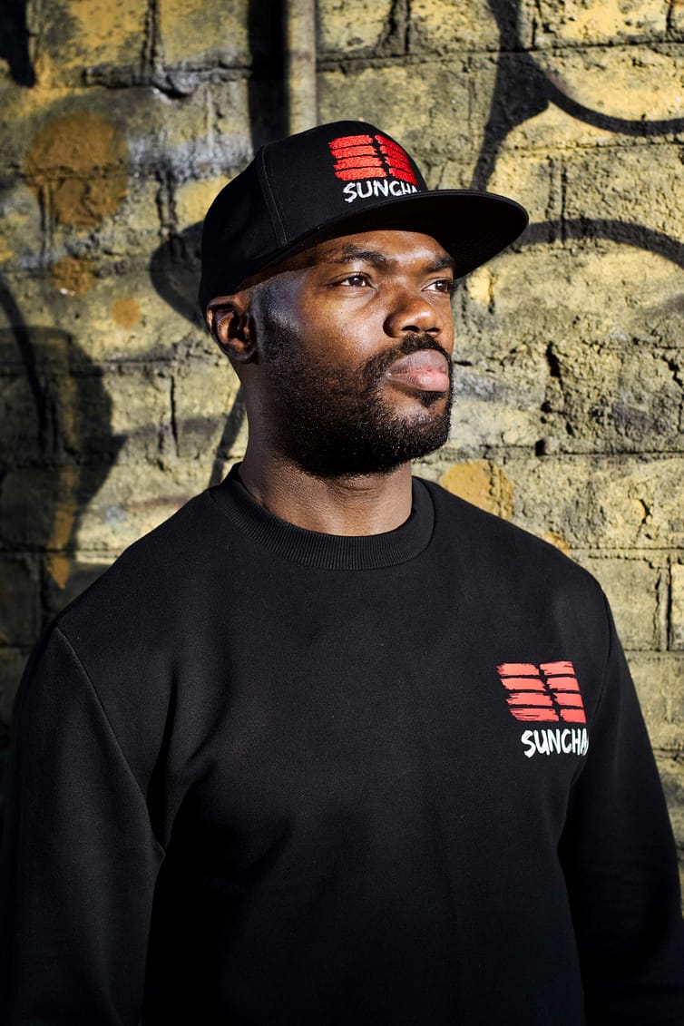 A man wearing a black sweatshirt and hat in front of a brick wall.