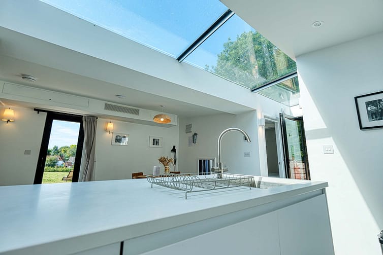 A modern kitchen with a large skylight, white countertops, and a view of a dining table and outdoor greenery through glass doors and windows— a lovely property shown off by expert photography by Ben Bowles