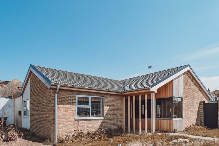 A single-story brick house with a gray tiled roof and wooden accents under a clear blue sky. The house features large windows and a small covered porch, construction property photography by Ben