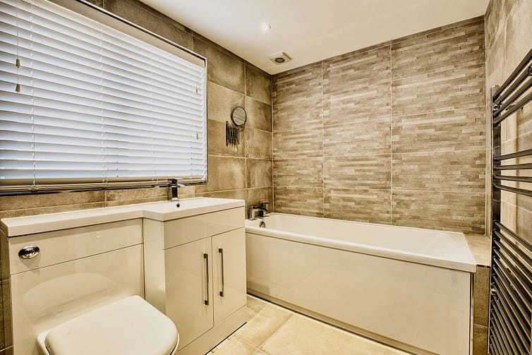 Modern bathroom with a tiled bath, white sink with chrome tap, and heated towel rail on a wall with stone tiles— great example of quality photography showcasing a new house
