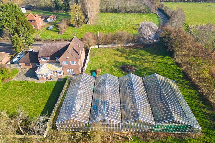 Aerial photograph of a rural area showcasing two houses with gardens, large greenhouses, a few parked cars, green fields, and local farmland — drone property photography by Ben Bowles
